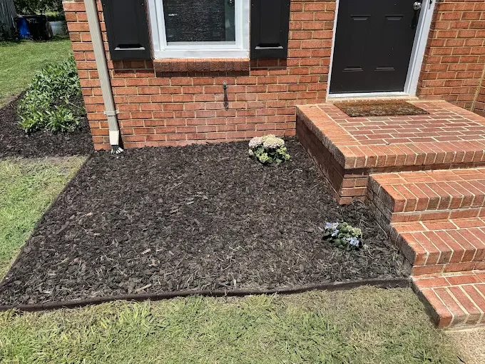 A front yard with freshly tilled soil, two small flower arrangements, brick stairs, and a brick house with a black door and window shutters.