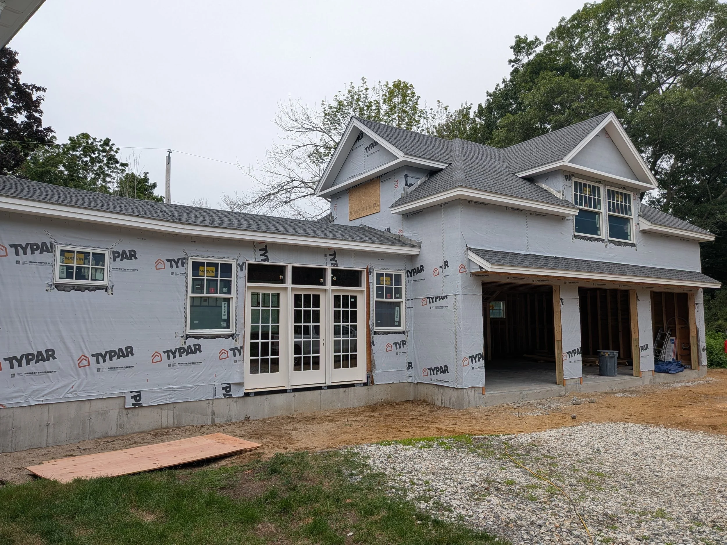 Front of a house under construction, with white weather-resistant building wrap, multiple windows installed, and a roof with dark shingles.