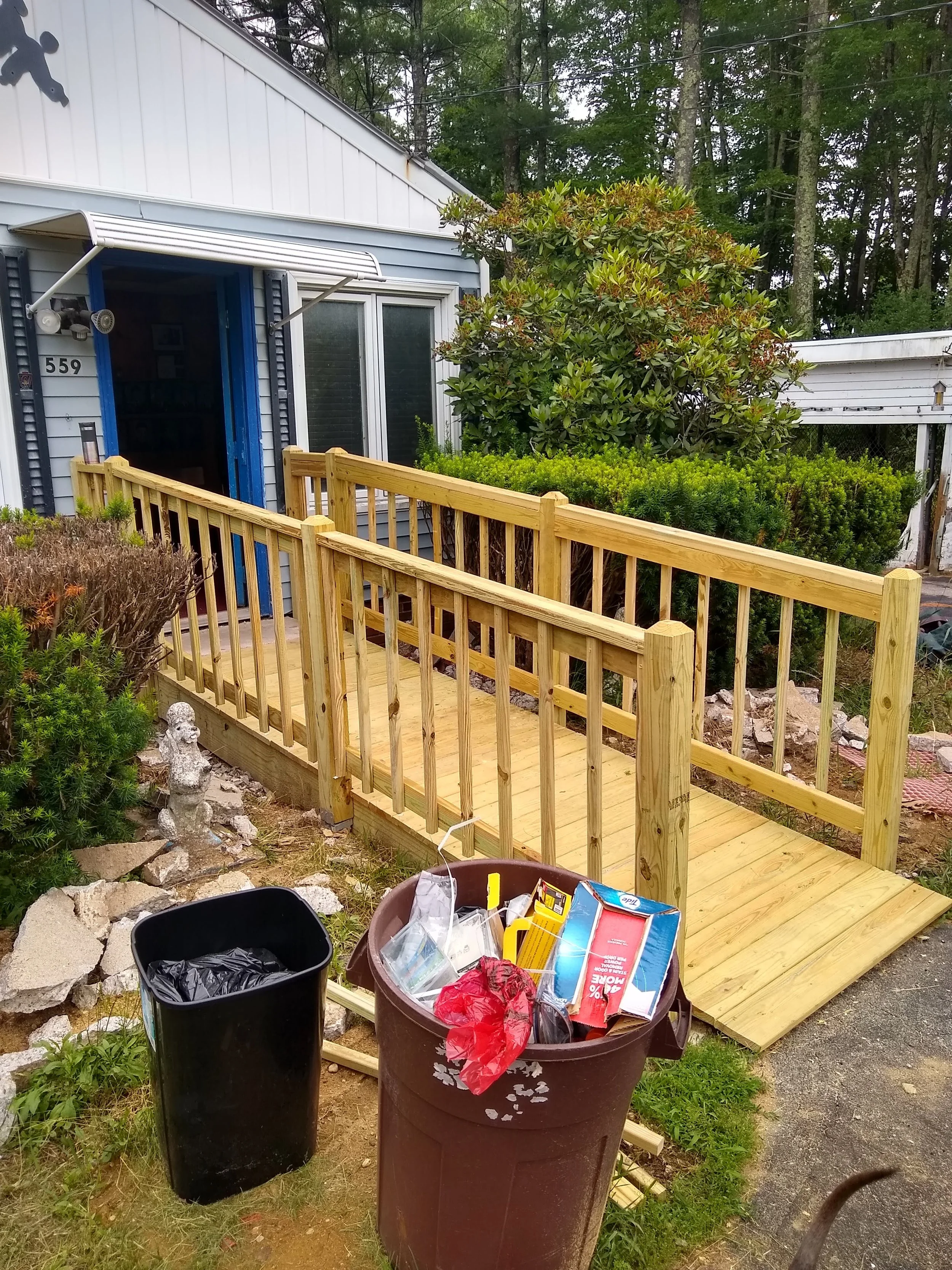 House with exterior wooden ramp under construction, surrounded by trash cans and plants, with trees in the background.