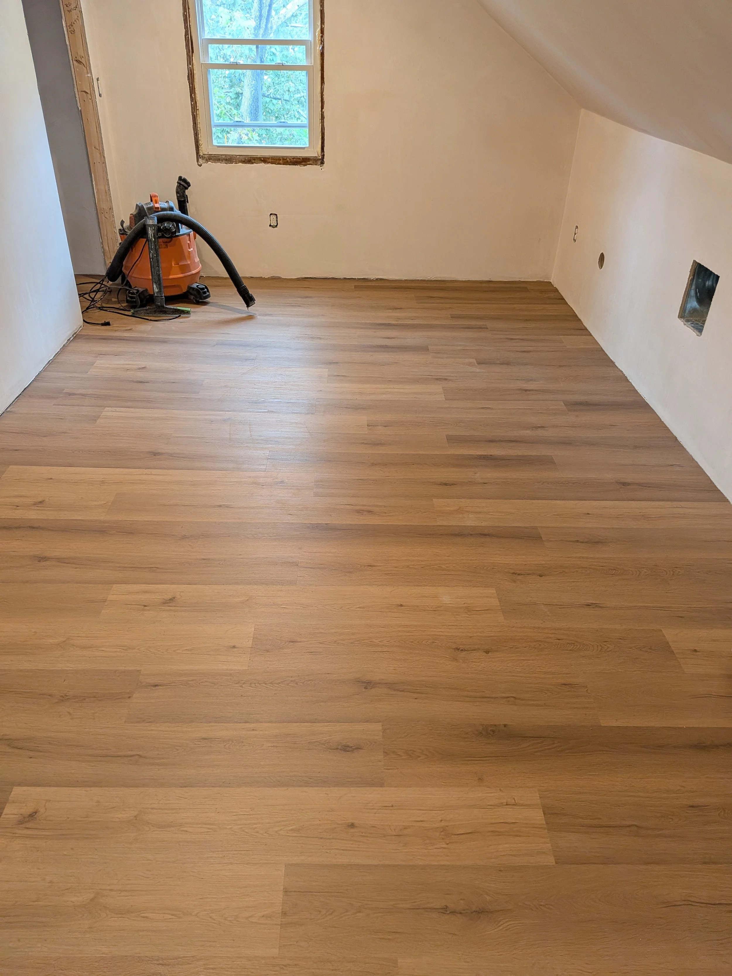 An empty room with wooden flooring, a window with a view of trees outside, and a vacuum cleaner placed near the wall.