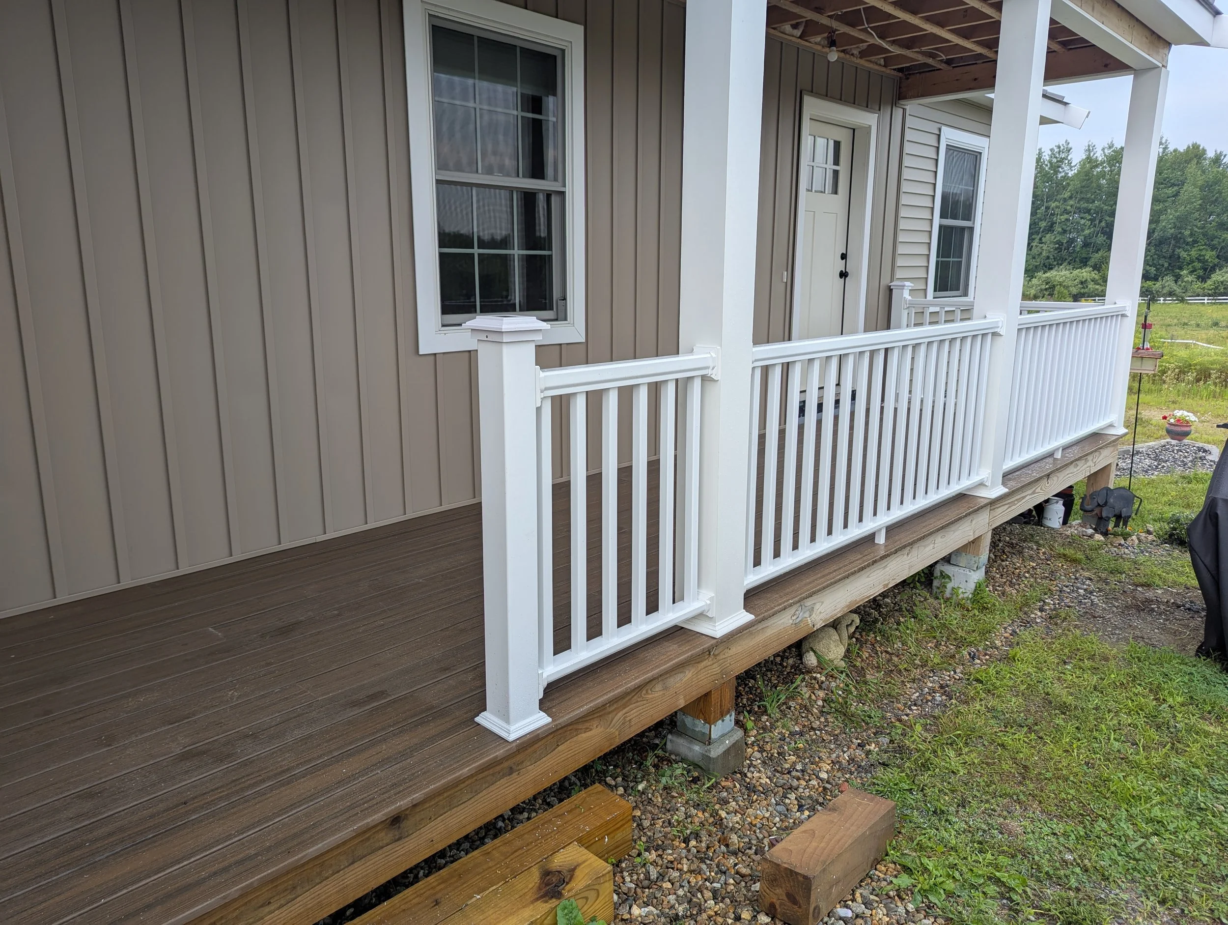 Back porch with weathered wooden deck covered in fallen autumn leaves, surrounded by grassy yard and leafless trees, with a wooded area in the background.