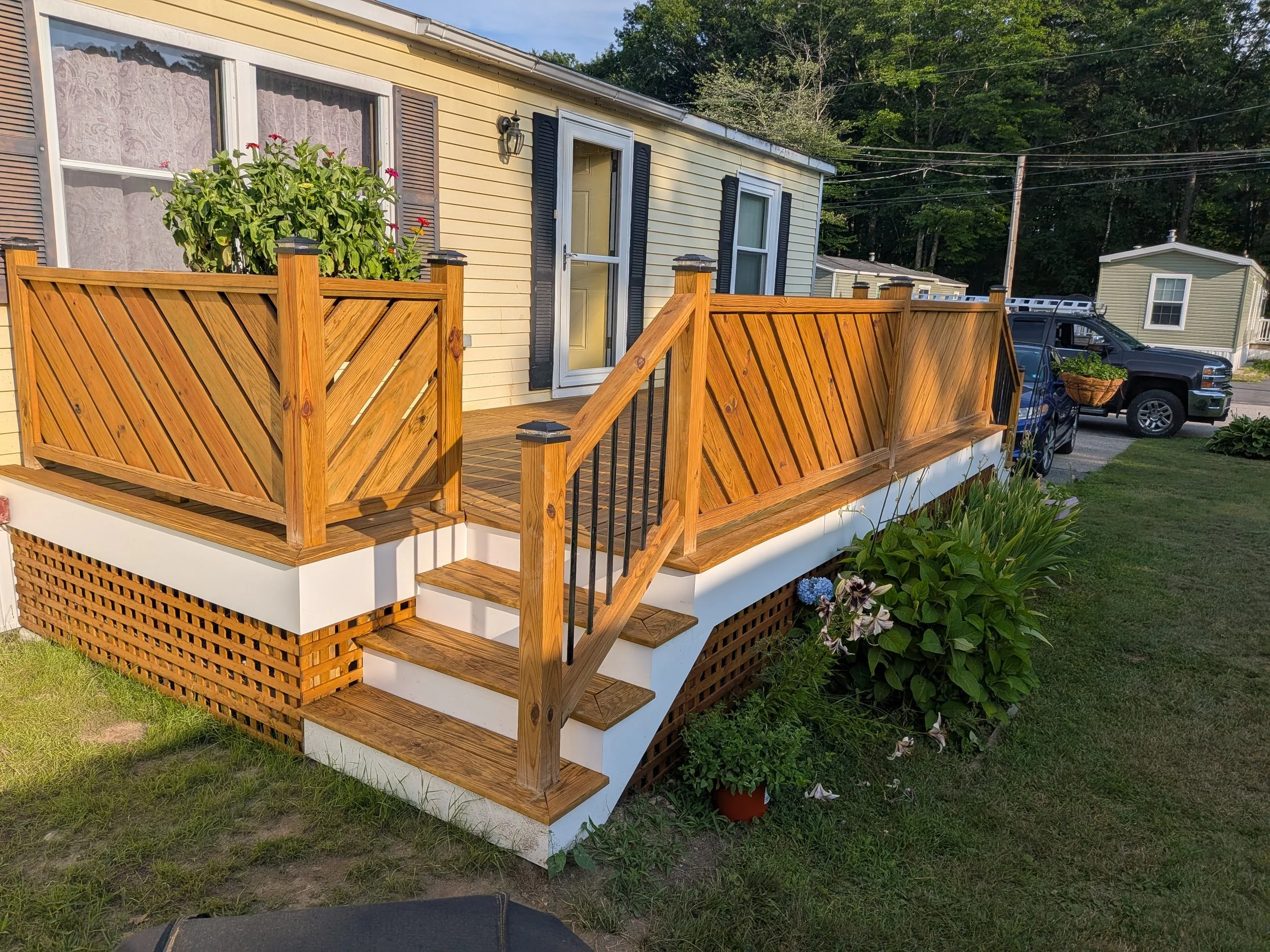 A house with a yellow exterior, black shutters, and a small wooden deck with stairs leading down. There is a flower pot with pink and purple flowers on the deck, and a vehicle parked nearby. The scene is outdoors with trees in the background.