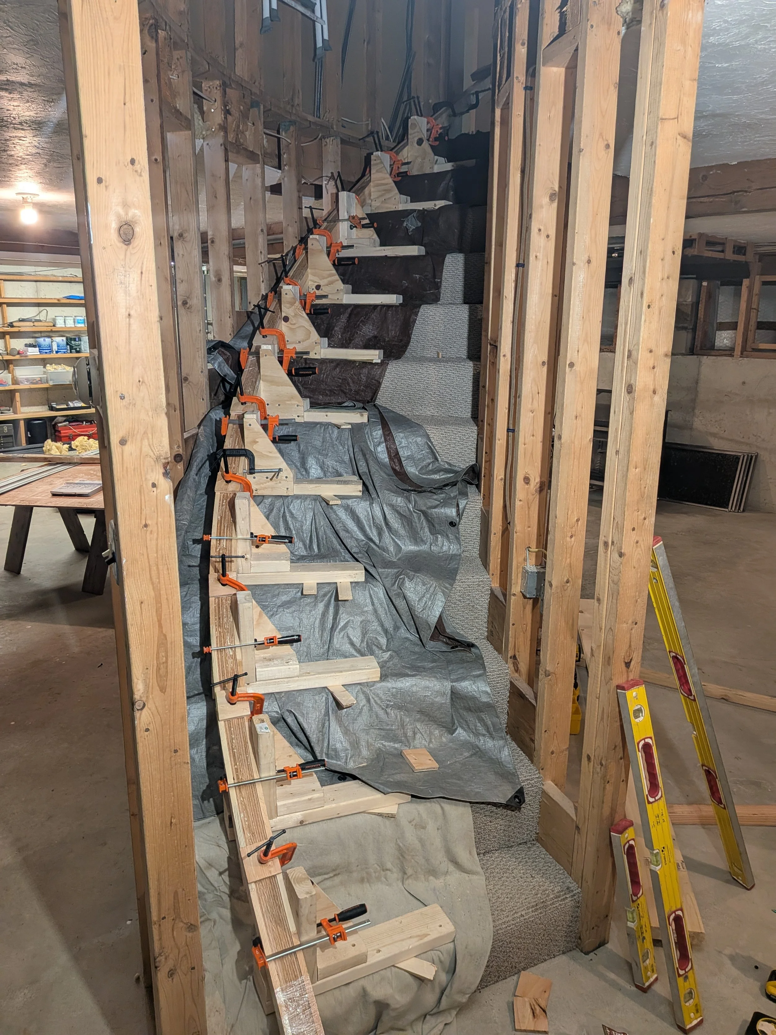 View of a house framing construction site with wooden studs, clamps holding attached wooden structures, a partially rolled gray carpet, and tools like a yellow ladder and a level.