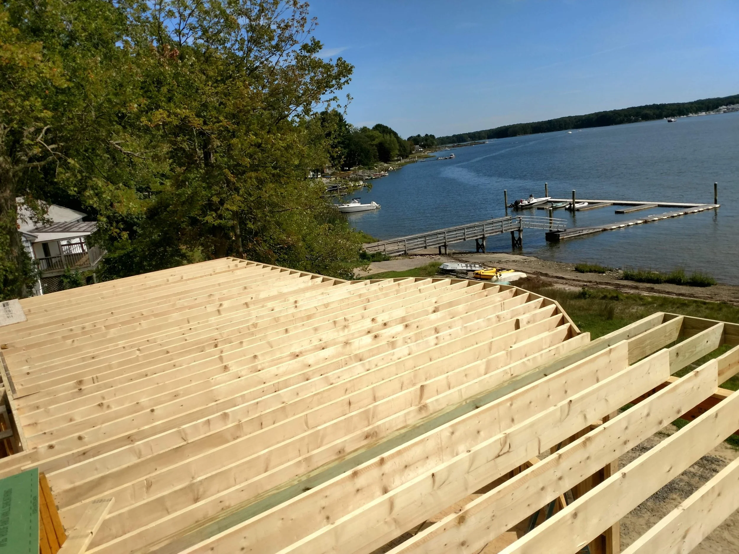 View of a lakeside with boats and docks, trees on the shoreline, and a partially constructed wooden deck in the foreground under a clear blue sky.
