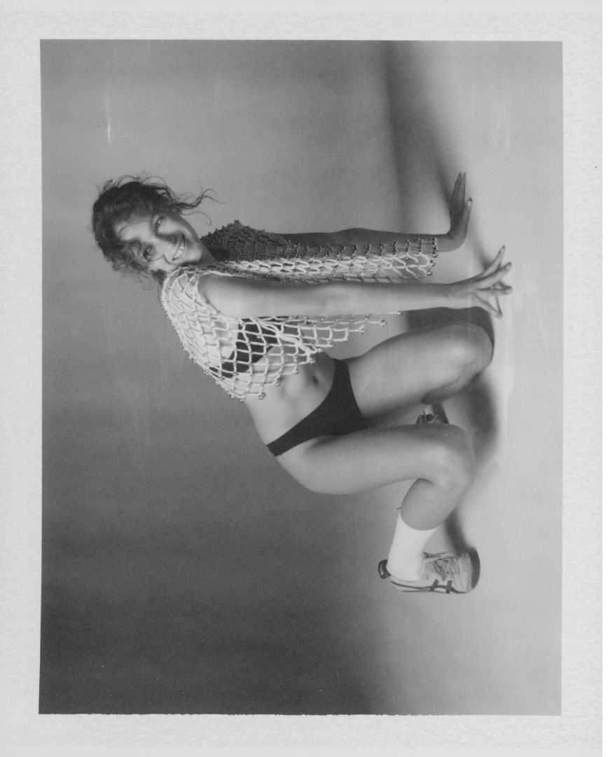Woman in black shorts and sneakers squatting against a plain wall, wearing a crochet top, smiling, black and white photo.