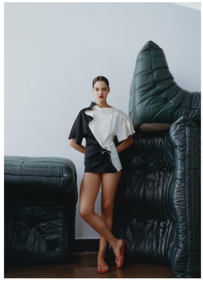 Corinne Murray in black and white Bottega Veneta with red sandals standing against a white wall, surrounded by oversized black leather furniture.