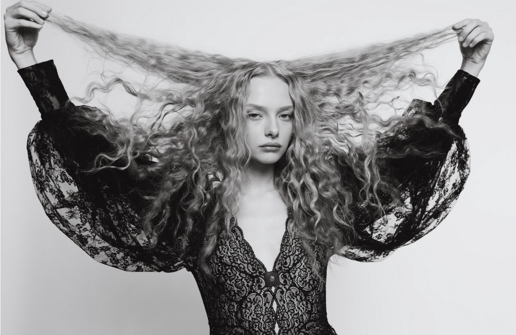 Amaiah Miller Black and white photo with long, curly hair, wearing a lace dress, holding her hair up with both hands.