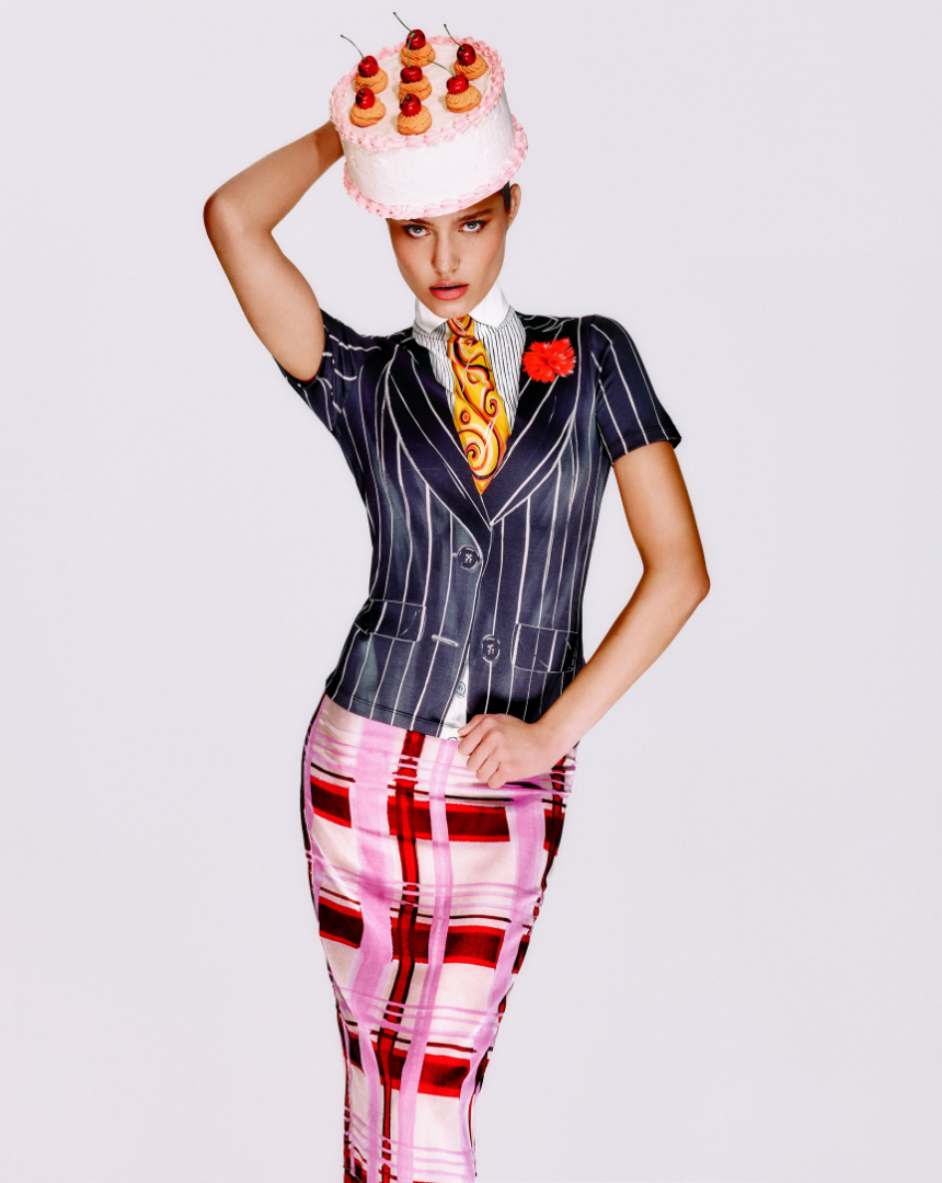 Woman in a Moschino striped blazer with a flower pin and colorful plaid skirt, balancing a cake with small pastries and cherries on her head.