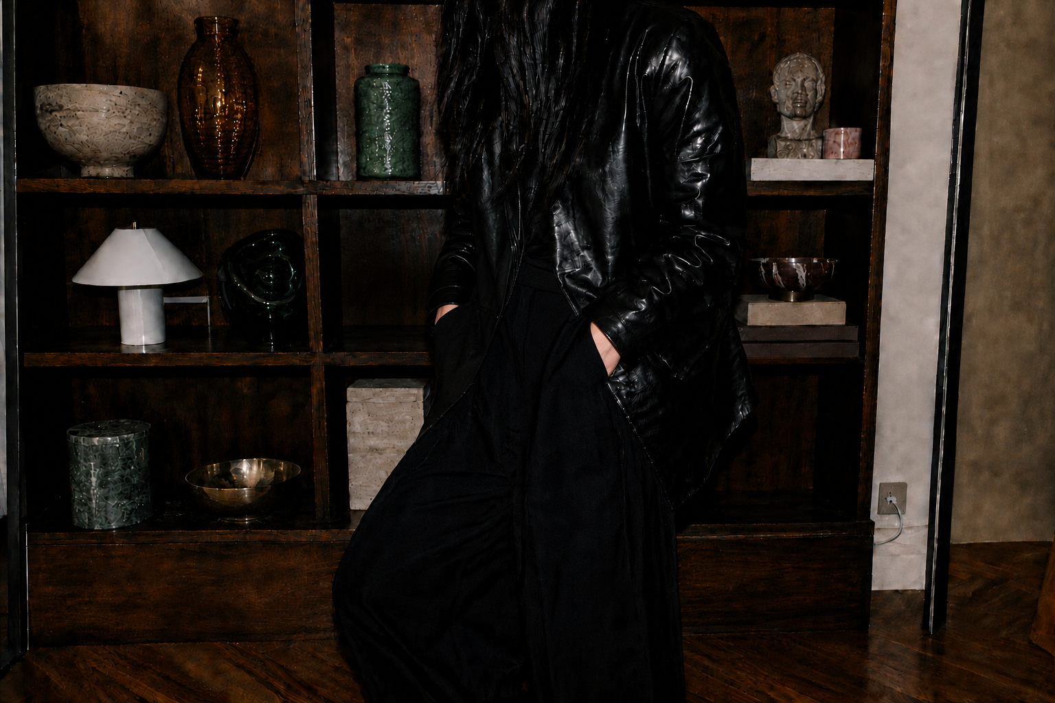 Veronica Lee Estes in a black leather jacket standing before a dark wood bookshelf filled with objects.