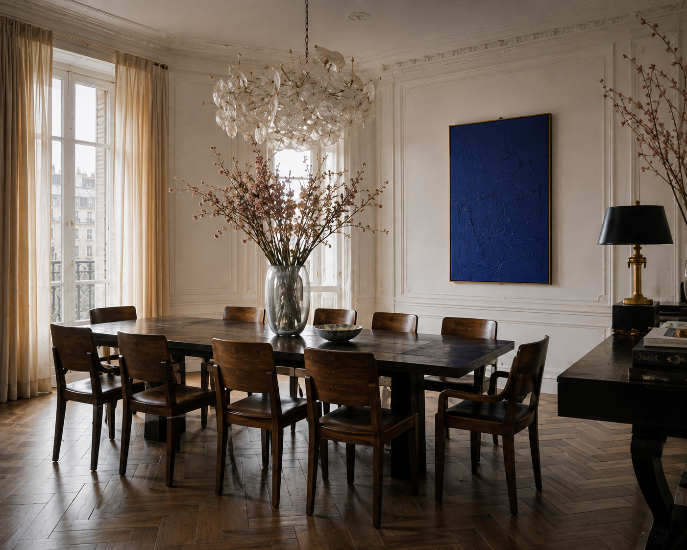 Formal dining room with dark wood table, sculptural chandelier, flowering branches, tall windows, and blue artwork.