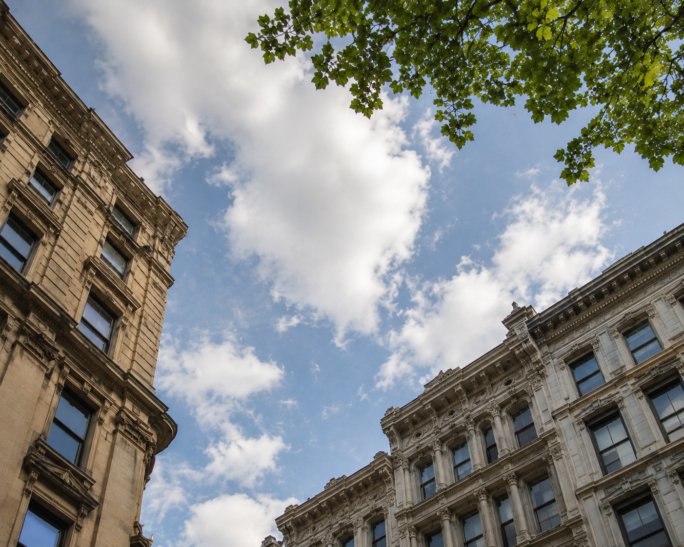 Blue sky and white clouds framed by ornate historic building facades and green leaves.