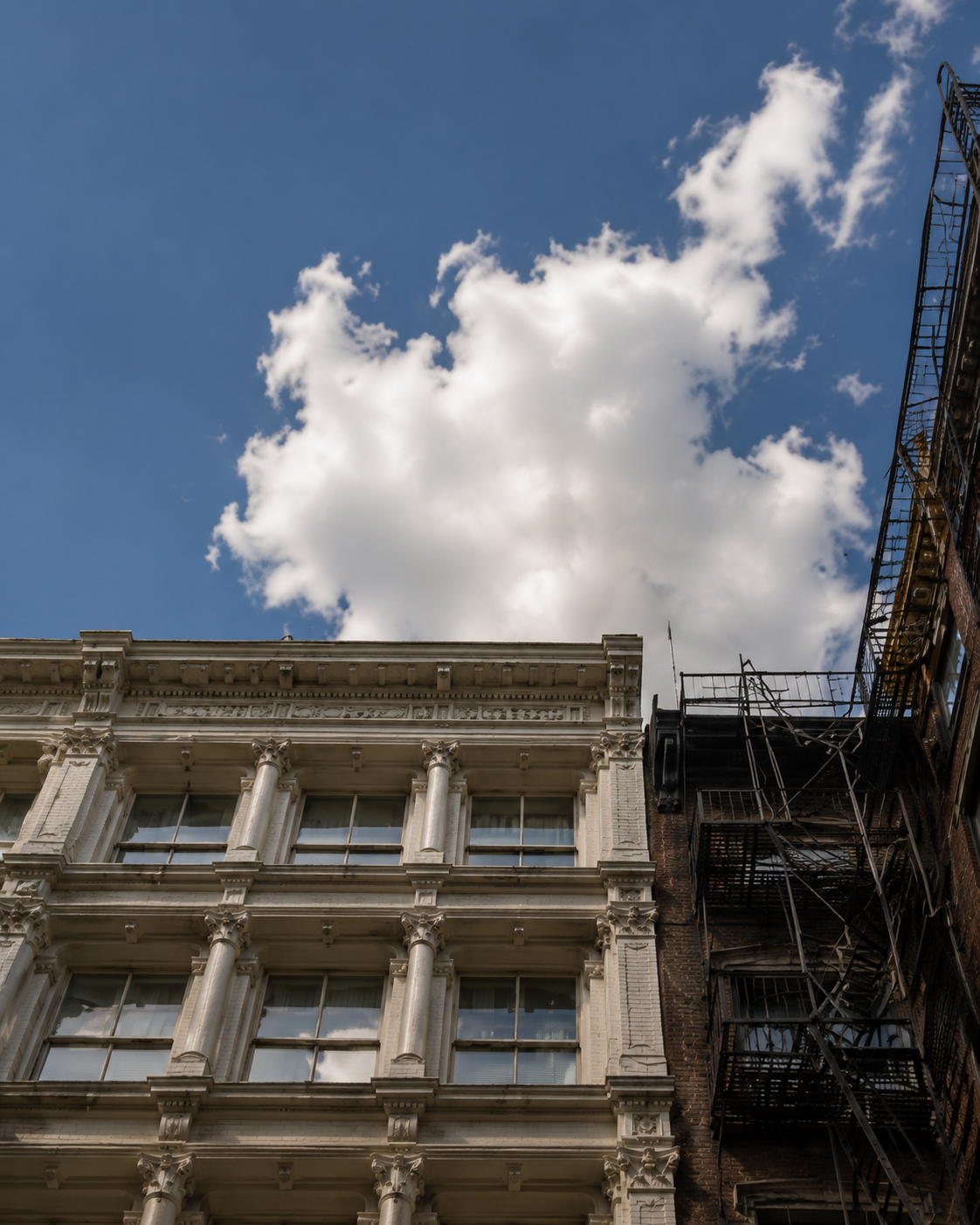 Large white cloud against blue sky above ornate historic facades and a metal fire escape.