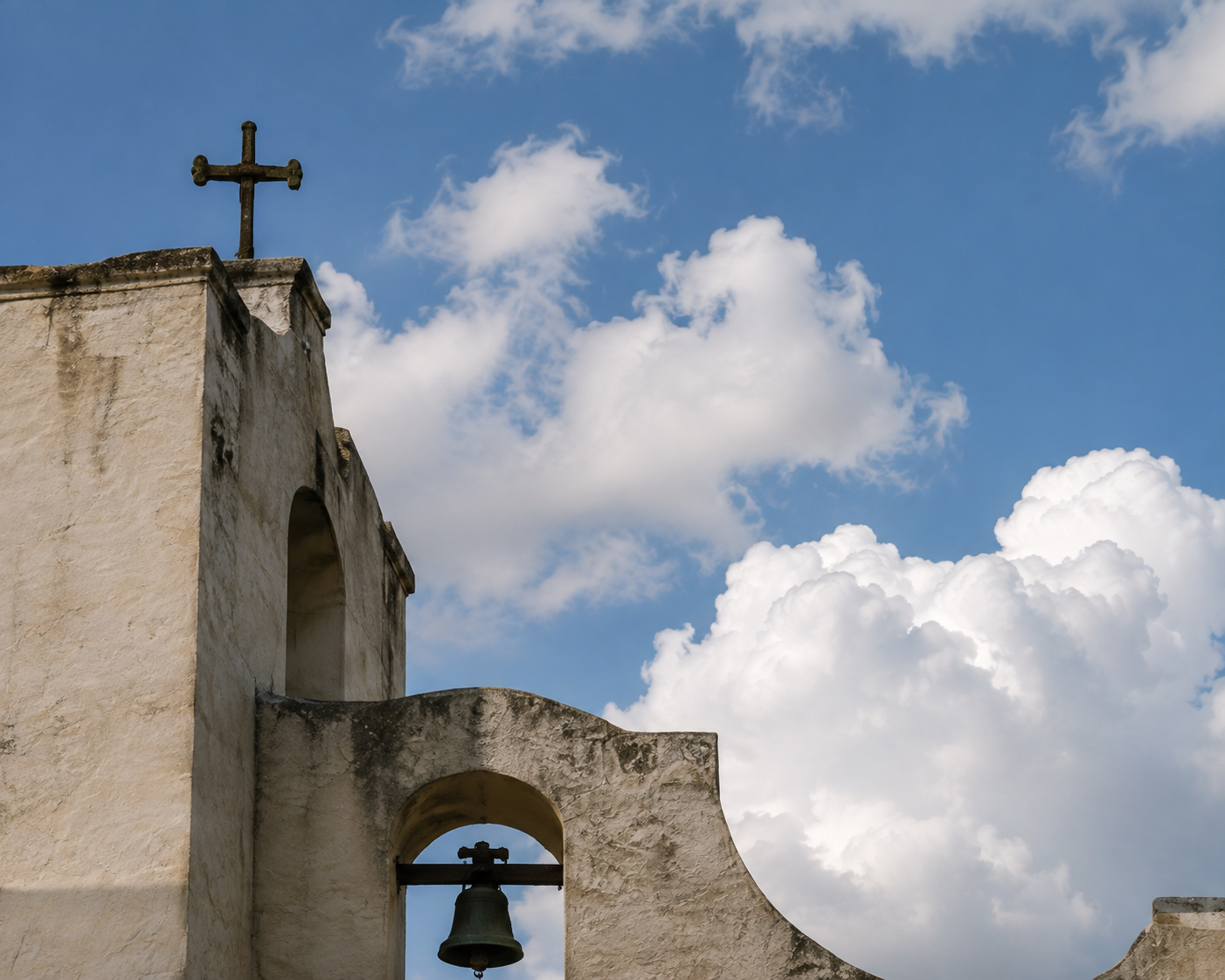 Weathered mission-style wall and bell opening beneath large white clouds.