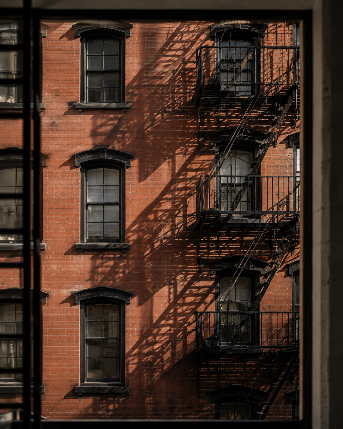 Red brick residential facade with black fire escapes casting geometric shadows.