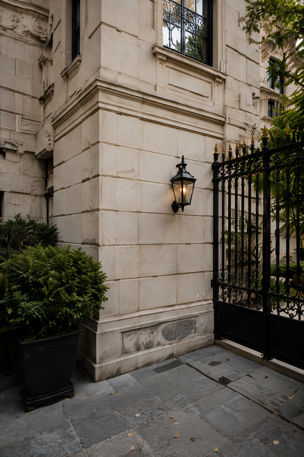 Limestone townhouse entrance with a black iron gate, wall lantern, and clipped greenery.