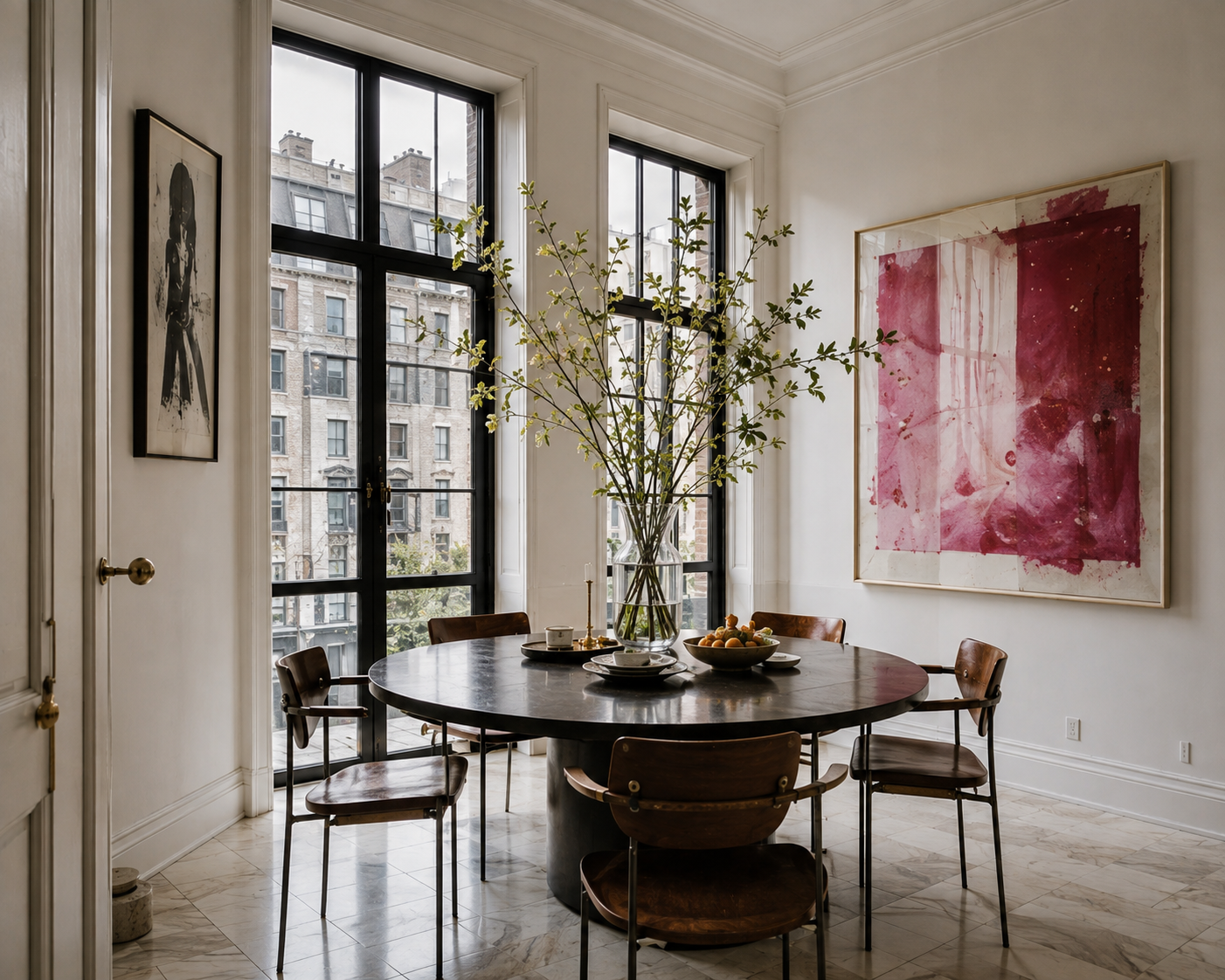 Dining area with black-framed windows, round table, wood chairs, leafy branches, and pink artwork.
