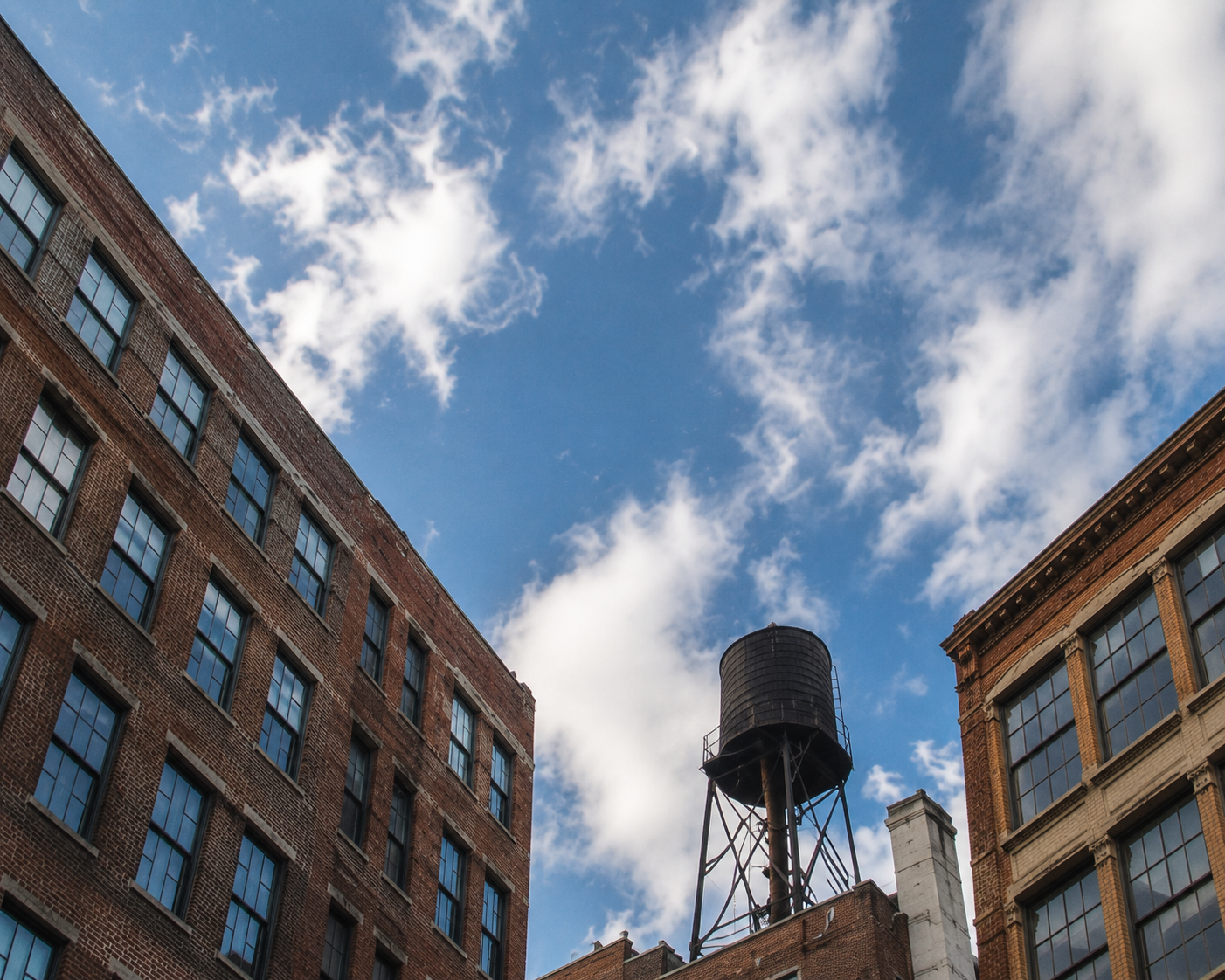 Rooftop water tower between brick buildings beneath a blue sky filled with white clouds.