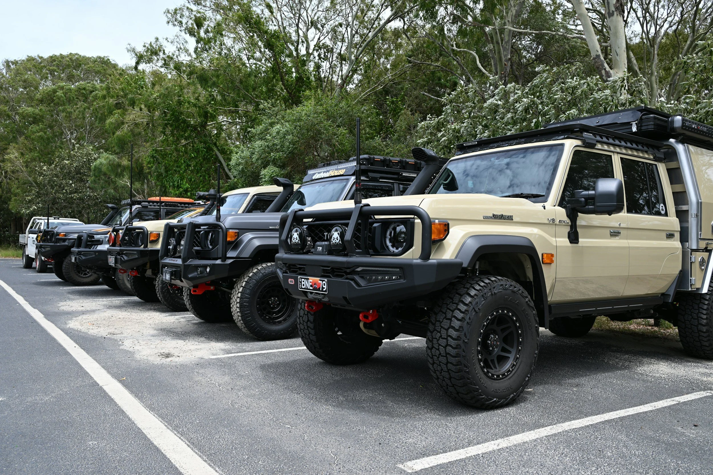 Line of off-road vehicles parked in a parking lot surrounded by tall green trees.