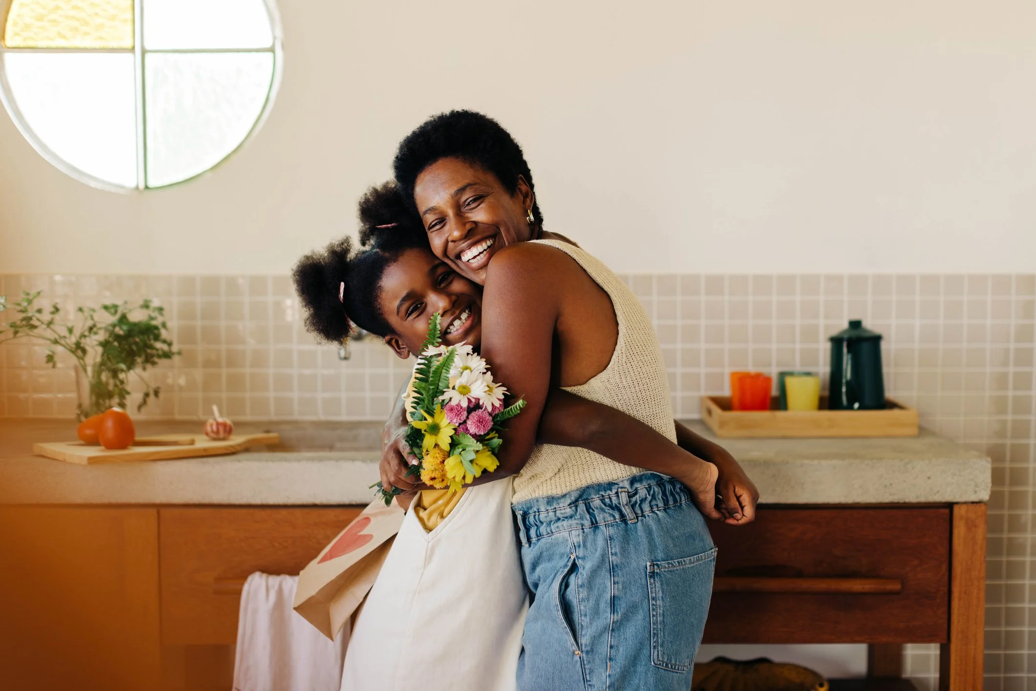 A woman and a young girl are hugging each other in a kitchen. The woman is holding a bouquet of flowers and smiling.