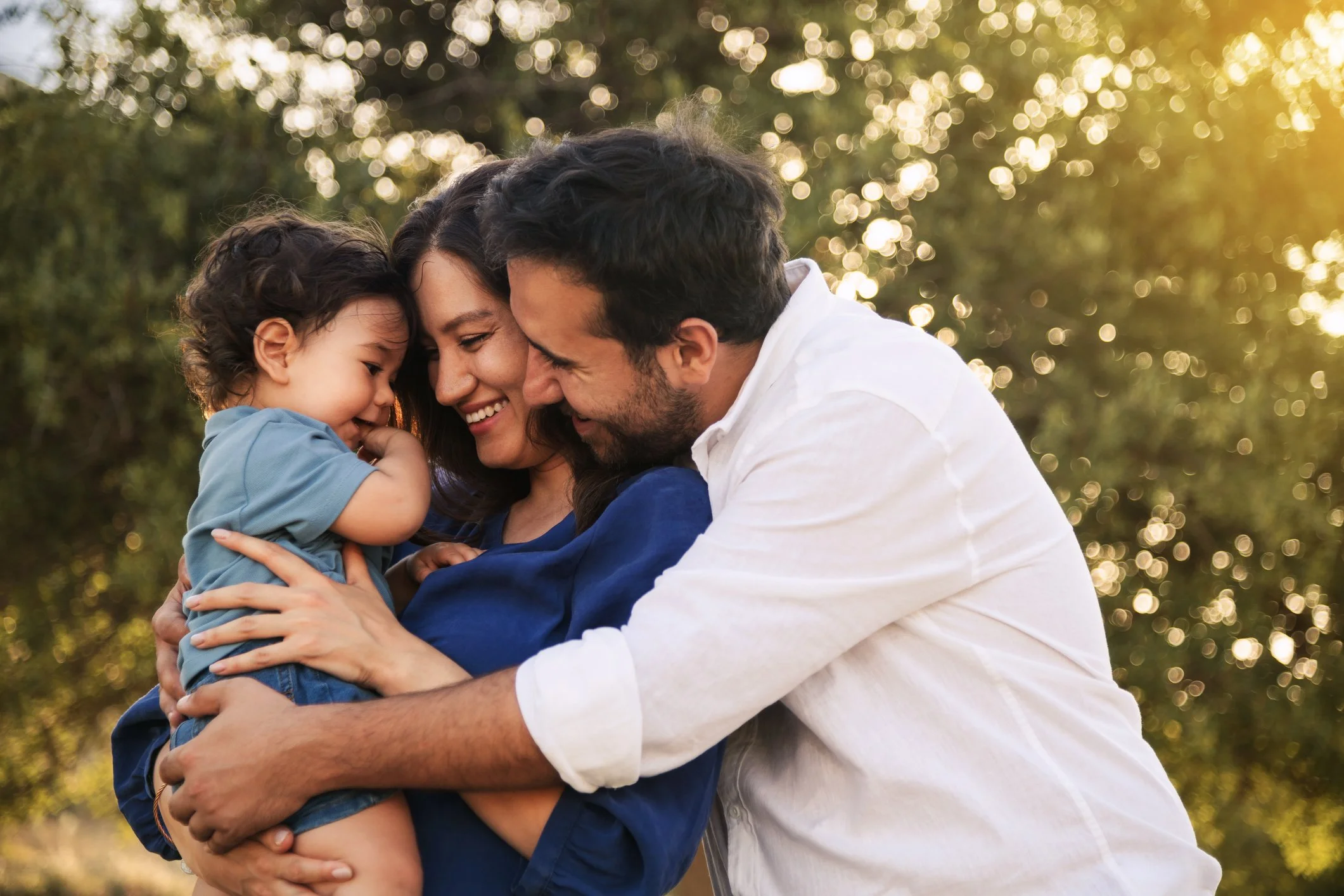 A happy family of three, including a mother, father, and son, sharing a hug outdoors during daytime with sunlight filtering through trees.