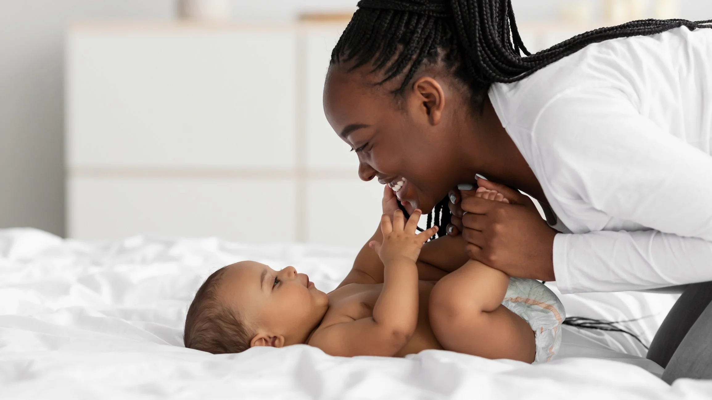 A woman and a baby playing and smiling on a bed, with the woman leaning over and holding the baby's hands, the baby looking up at her.