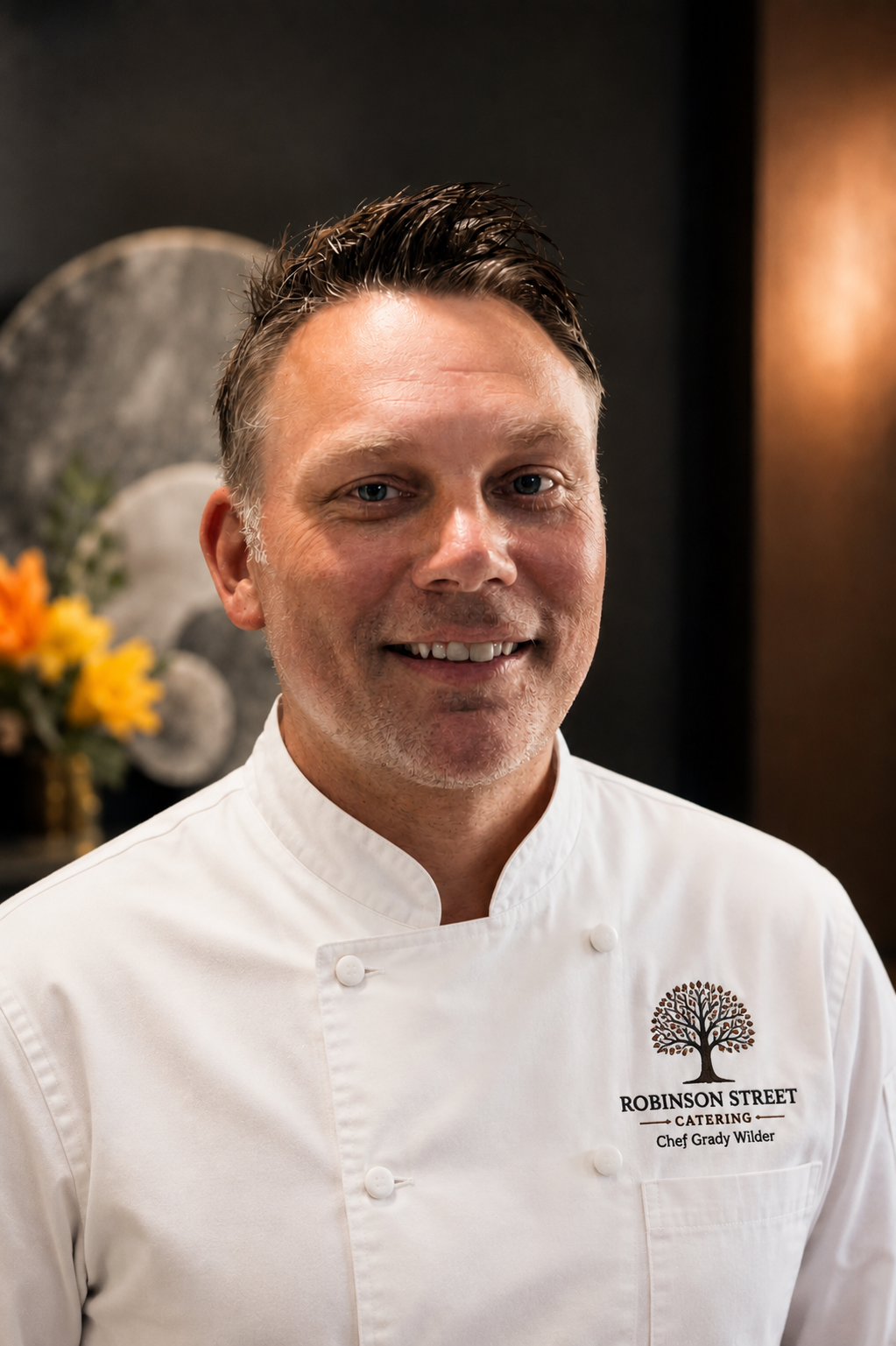Portrait of Chef Grady Wilder in white uniform with 'Robinson Street Catering' logo, smiling in a professional kitchen or dining area.