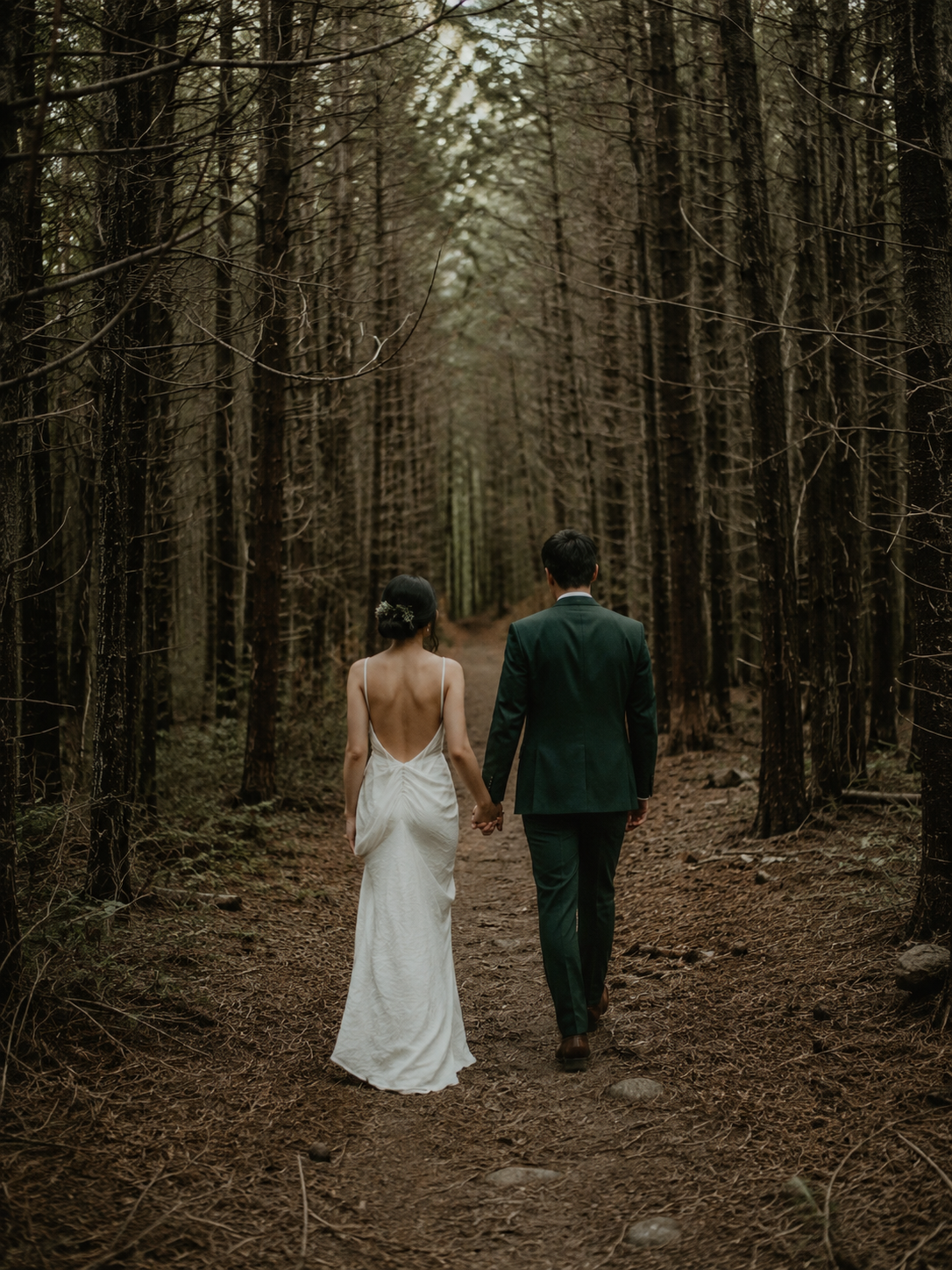 A bride and groom holding hands while walking through a wooded forest path during daytime.