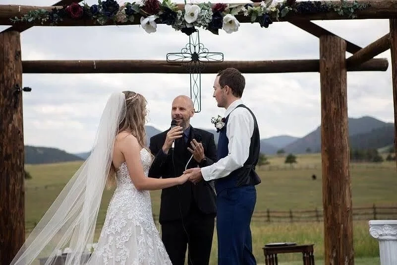 Bride and groom holding hands during an outdoor wedding ceremony under a wooden arch decorated with flowers, officiant smiling and holding a microphone, scenic countryside background with mountains and cloudy sky.
