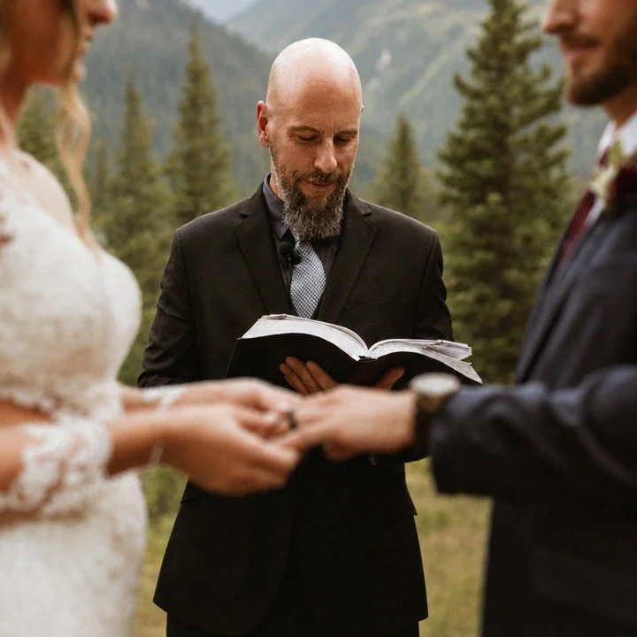 A priest officiating a wedding ceremony outdoors with trees and mountains in the background, holding an open book, with a bride and groom exchanging rings.