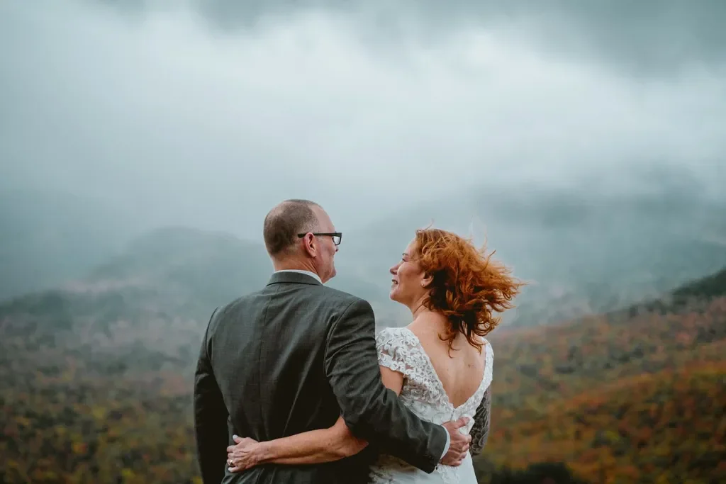 A couple dressed in wedding attire, embracing and smiling at each other outdoors with mountains and clouds in the background.