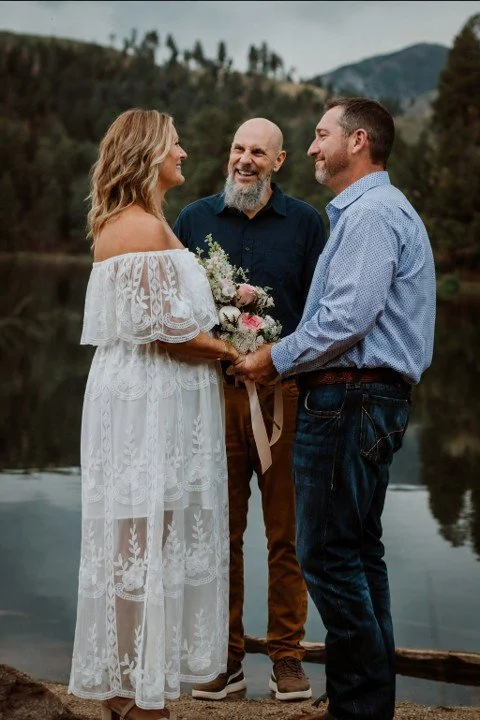 A woman in a white lace dress and a man in a blue shirt holding hands and facing each other, exchanging vows during a wedding ceremony by a lake with a man officiant and a scenic mountain background.