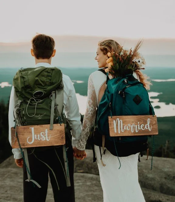 A newly married couple holding hands and looking at each other outdoors at sunset, each wearing a backpack with a wooden sign that reads "Just" and "Married".