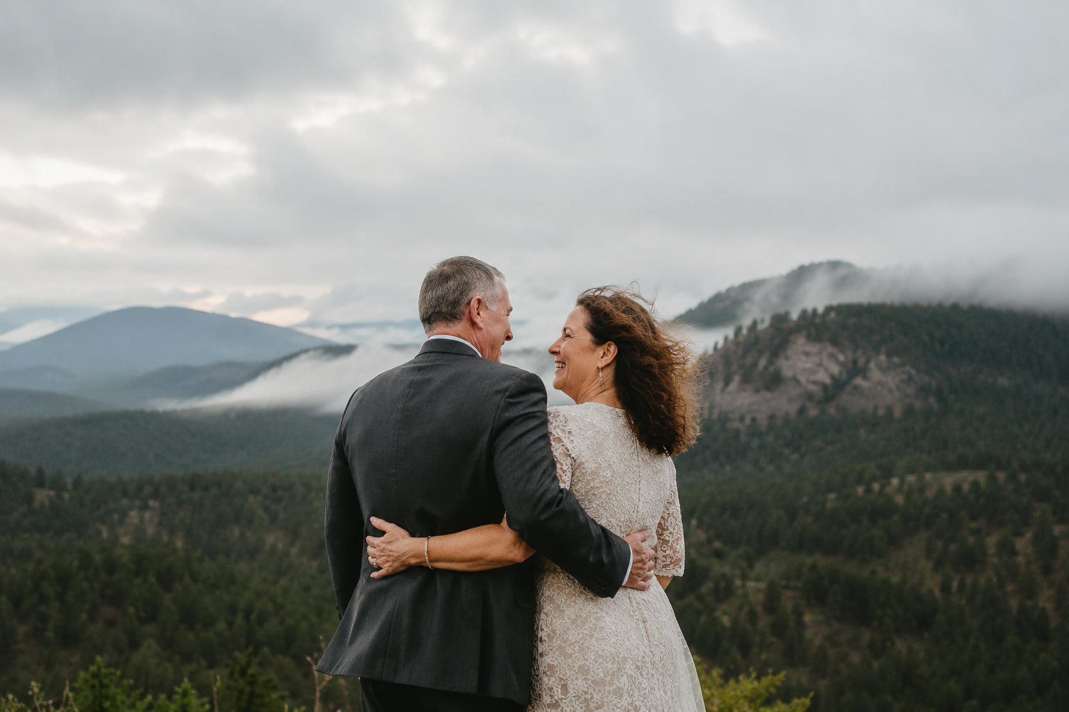 A couple dressed in wedding attire standing outdoors in front of a mountain landscape with clouds and fog, smiling and embracing each other.