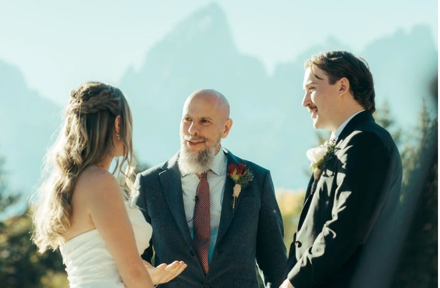 A couple getting married outdoors with an officiant, surrounded by mountains and trees, during daytime.