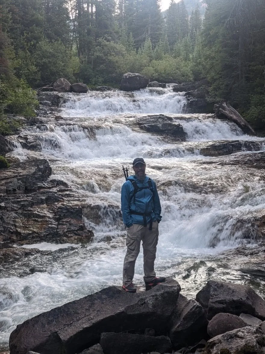 A person in outdoor hiking gear stands on rocks in front of a flowing mountain stream surrounded by dense green trees.