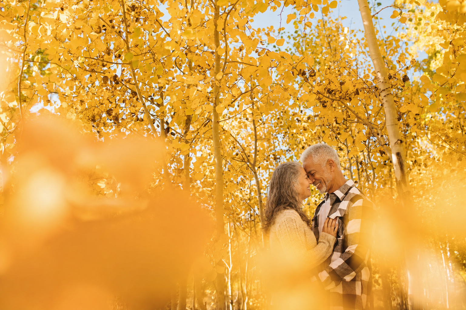 An elderly couple smiling and touching foreheads in a forest with vibrant yellow autumn leaves.