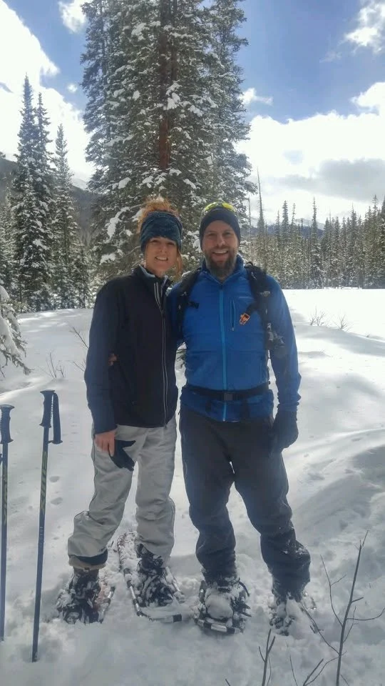 Two people standing in snow with winter gear, surrounded by snow-covered trees and mountains, on a bright day.
