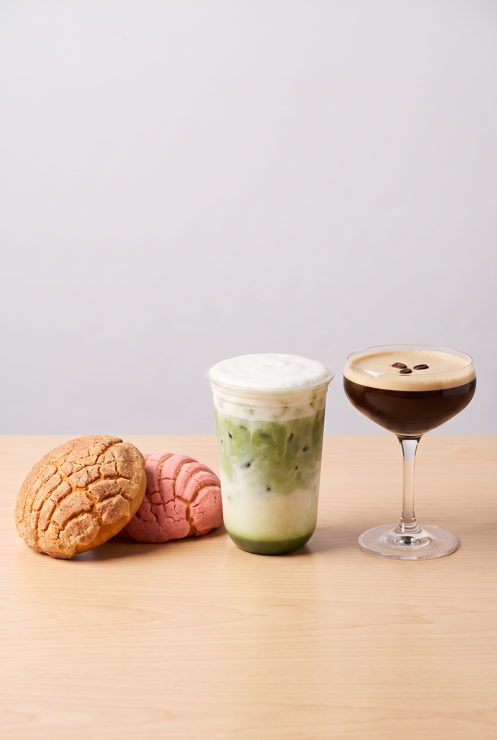 Oatmeal cookies and pink cookies on a wooden surface, a green and white iced drink in a glass, and a cappuccino in a glass cup with coffee beans on top.