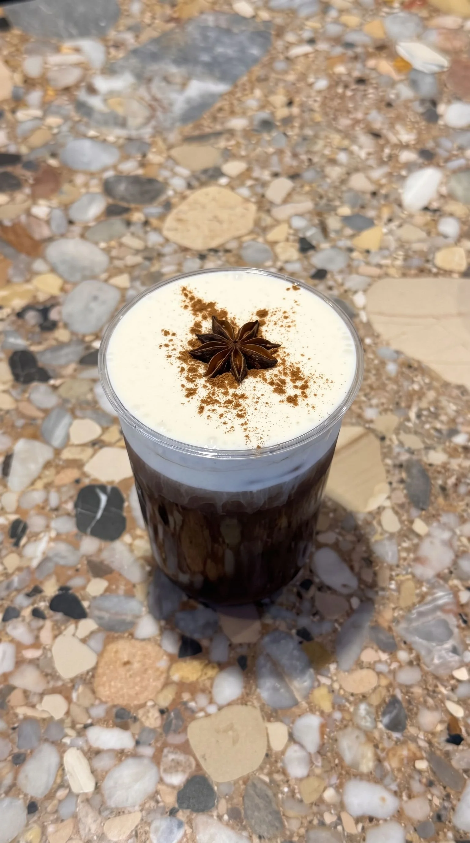 A glass of iced coffee topped with whipped cream, cinnamon, and a star anise spice, on a speckled stone countertop.