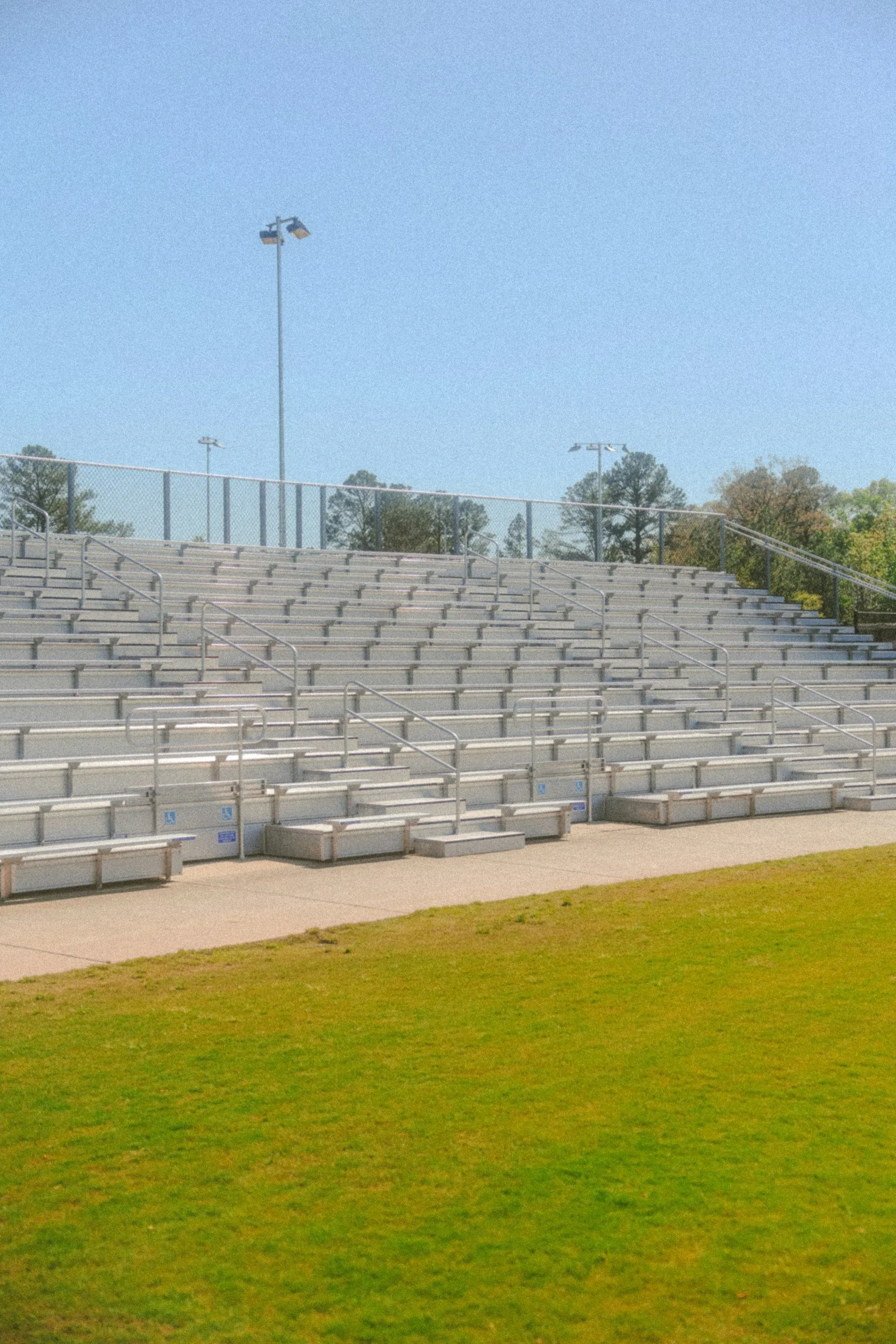 Empty outdoor stadium bleachers with a chain-link fence in the background, surrounded by trees and a clear blue sky.