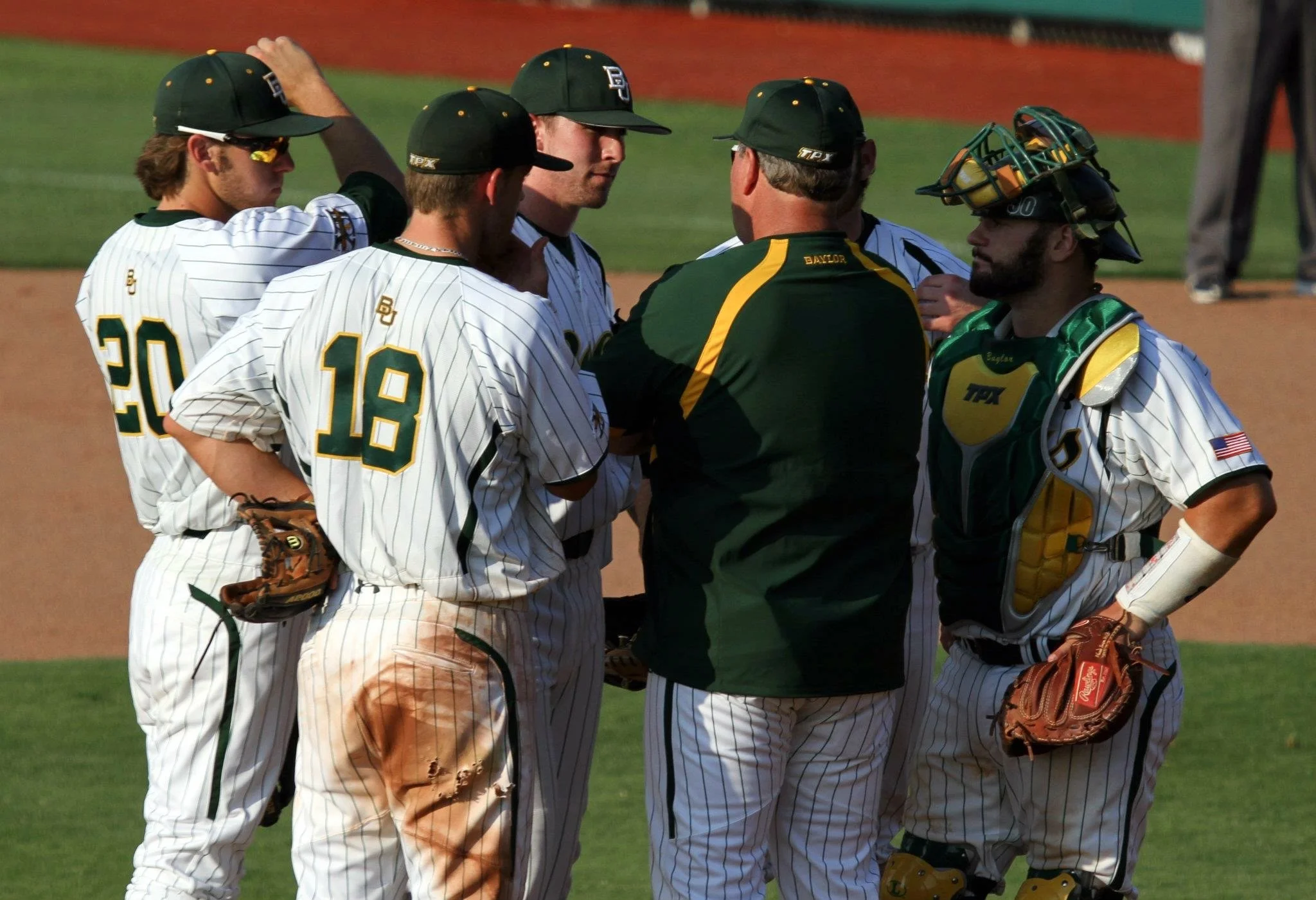 A group of six baseball players and a coach in a huddle on a baseball field. The players are in white pinstriped uniforms with green and yellow accents, some wearing caps and one wearing catcher's gear. The coach, dressed in a green and yellow shirt, is talking to the players.