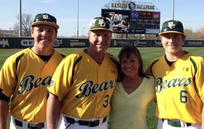 Four people, two young men, one middle-aged man, and one woman, standing on a baseball field wearing yellow jerseys with 'Bears' written on them. The two young men and middle-aged man are wearing baseball caps, and all are smiling. In the background, there is a scoreboard and stadium.