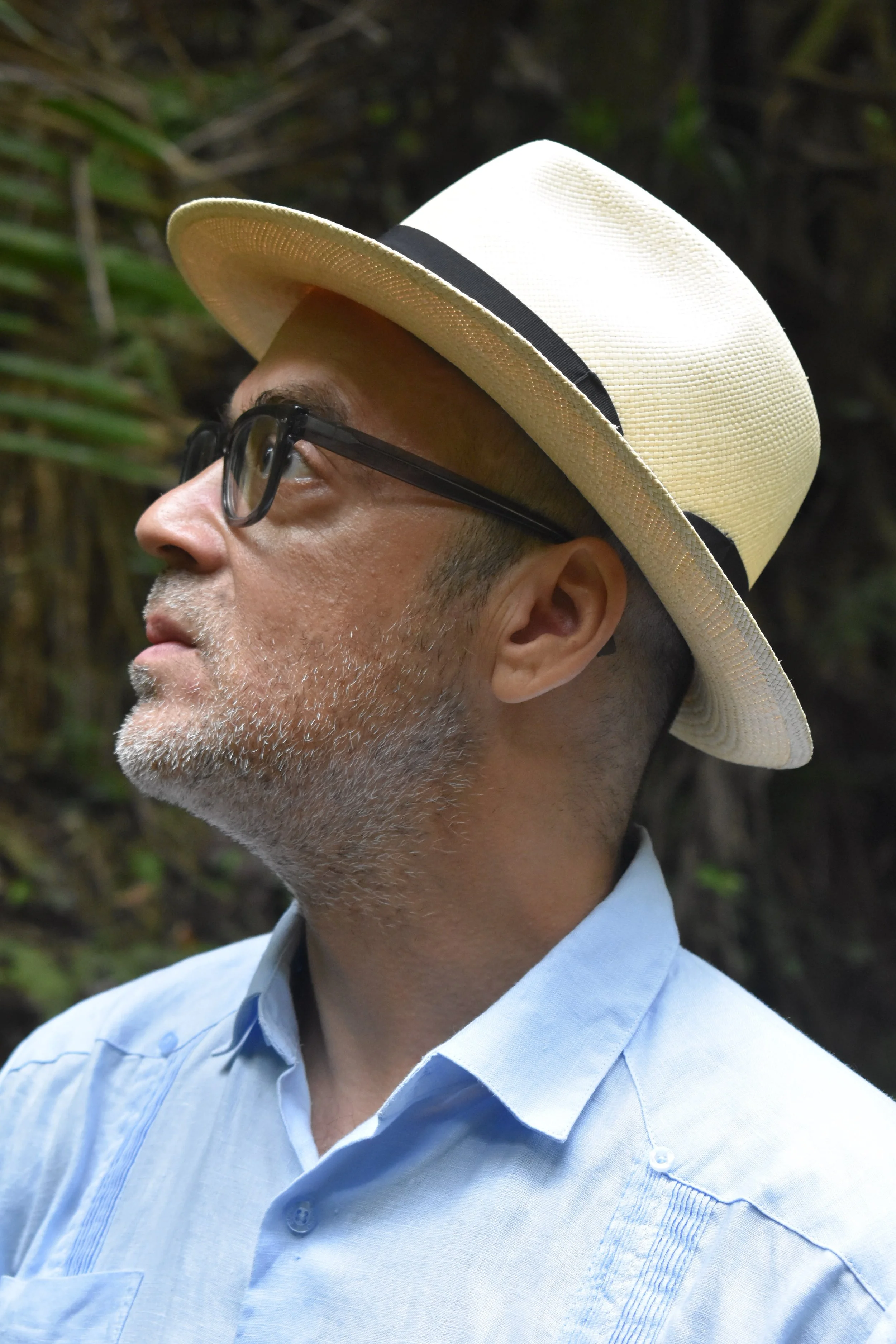 A man wearing glasses, a light blue shirt, a white straw hat with a black band, and looking to his left outdoors with a background of greenery.