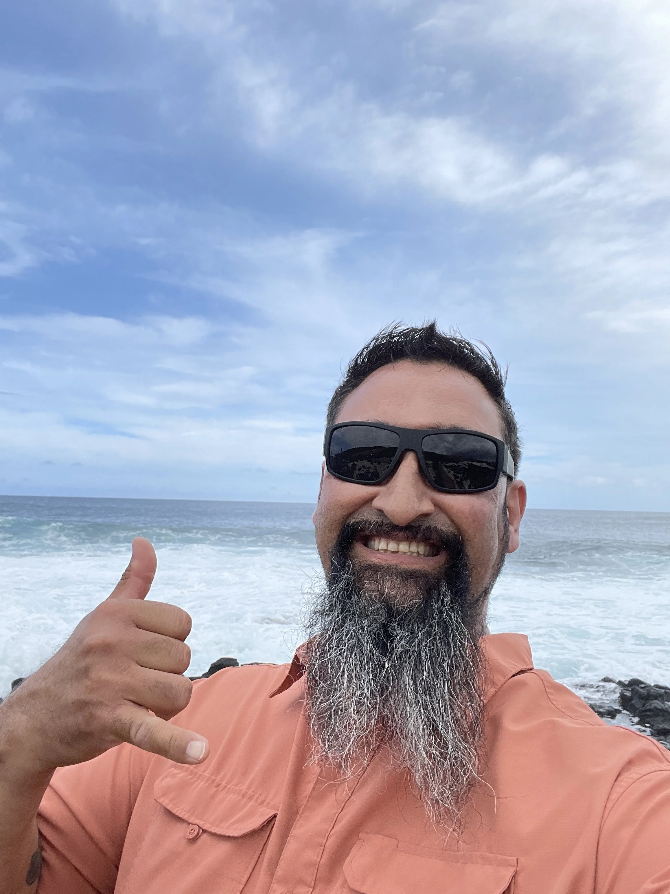 A man with a beard, black sunglasses, and a peach-colored shirt taking a selfie at the beach, smiling and giving a thumbs-up gesture with the ocean and sky in the background.