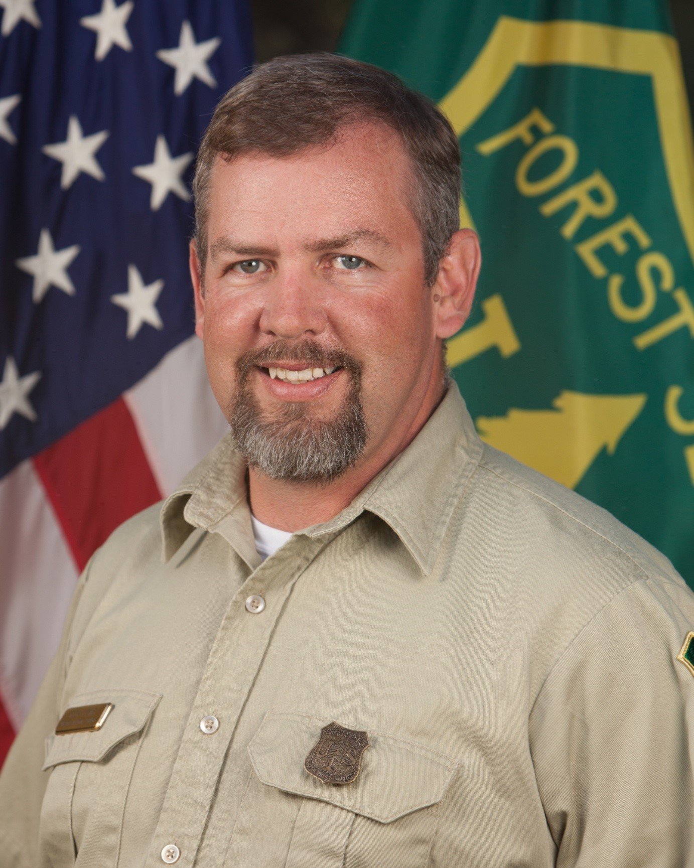 A man with gray hair and a beard wearing a beige uniform with a badge in front, standing in front of American and green flags.