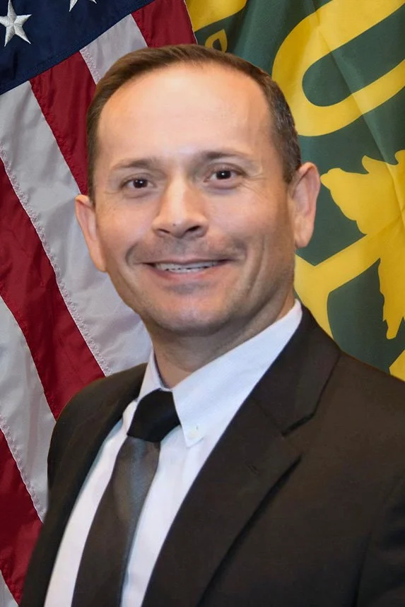 A man in a black suit, white shirt, and black tie smiling, standing in front of an American flag and a green flag with a yellow emblem.