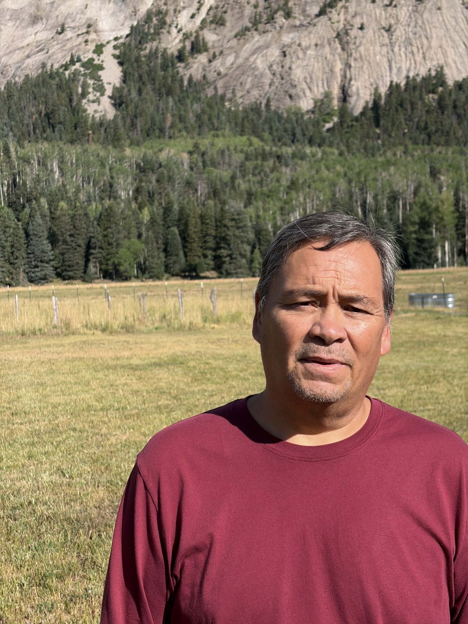 A man standing outdoors, wearing a maroon long-sleeve shirt, with a background of trees, mountains, and a grassy field.