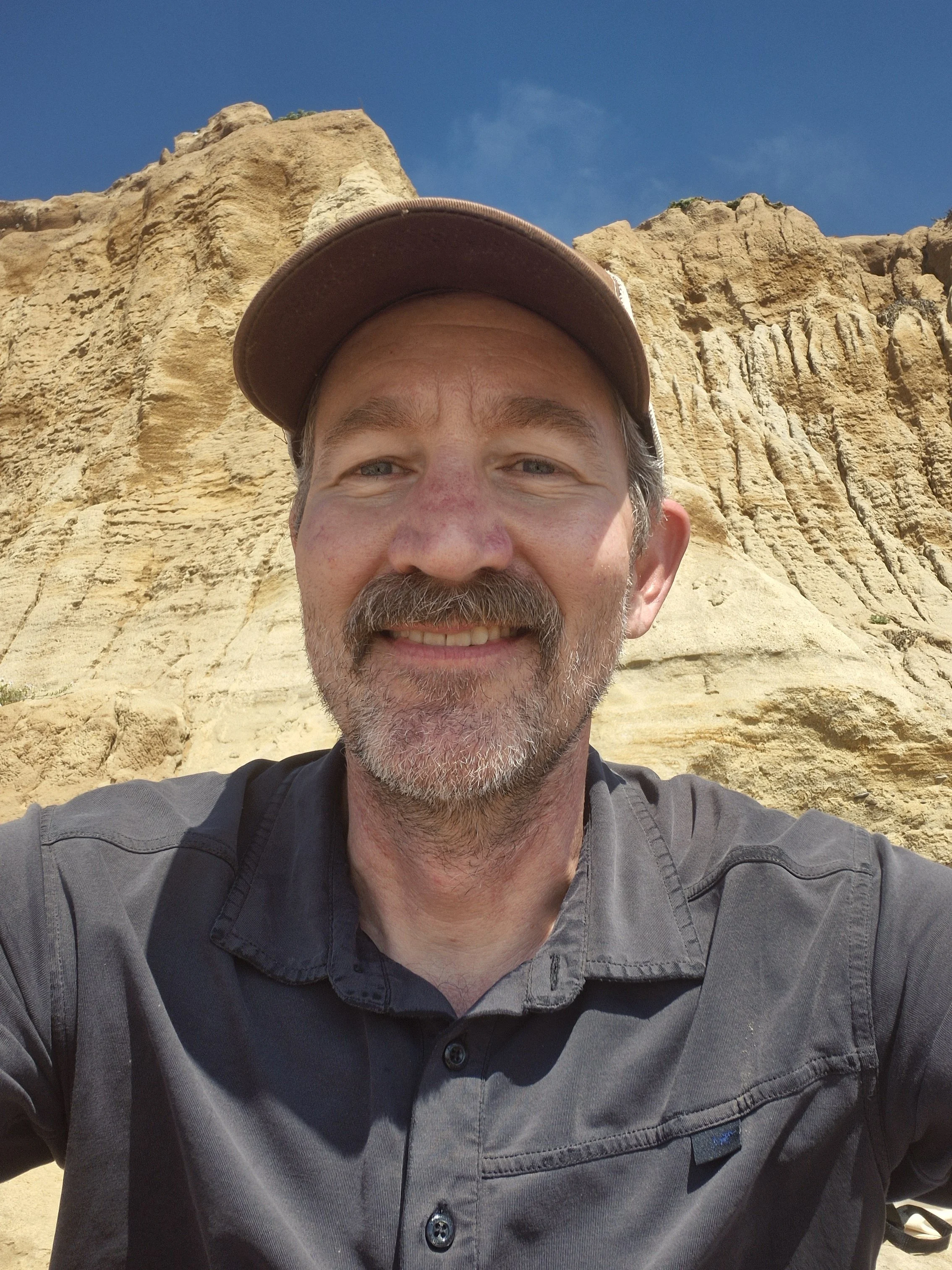 Close-up of a smiling man with a beard, wearing a brown baseball cap and a gray button-up shirt, standing in front of rocky cliffs and a clear blue sky.