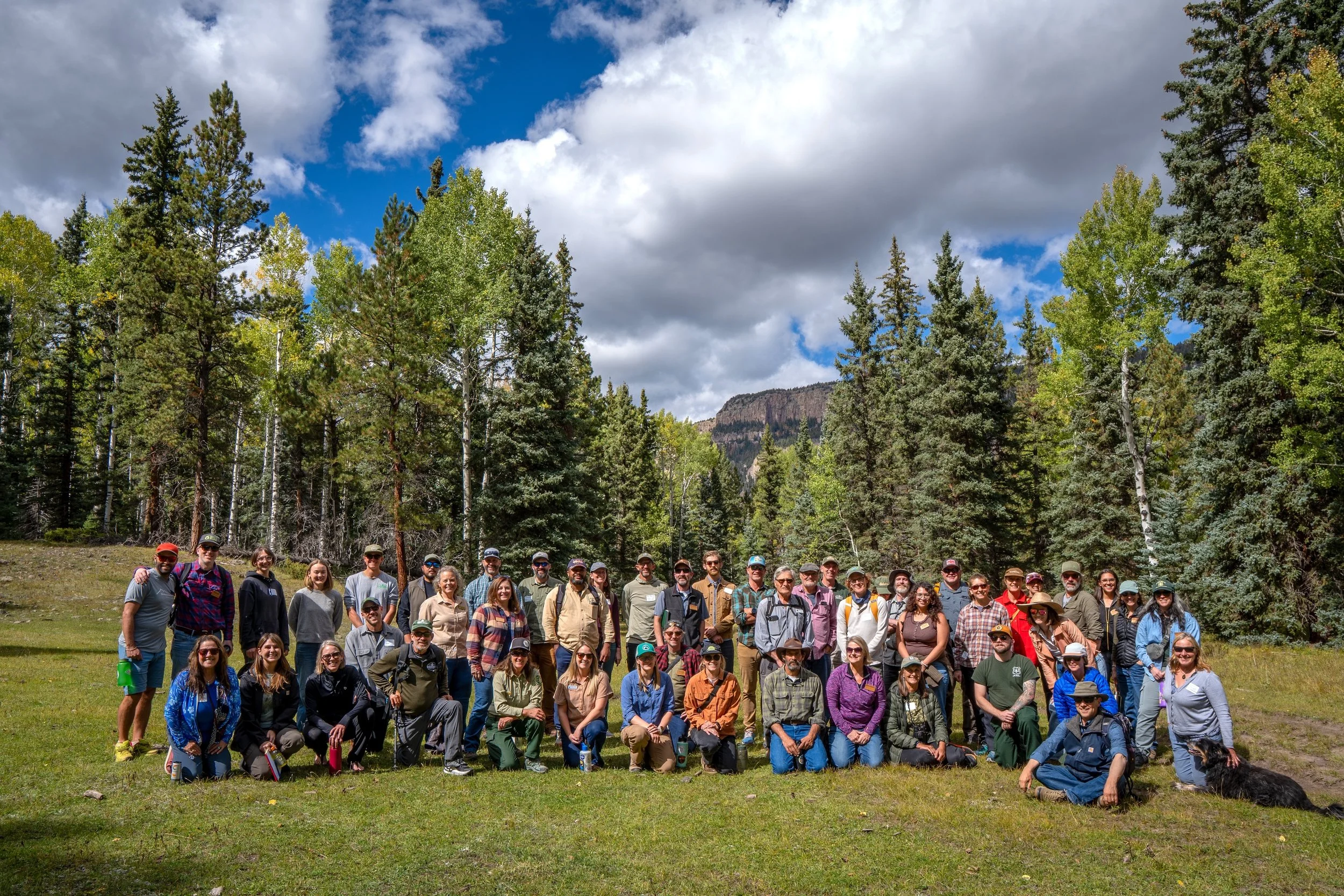 A large group of people outdoors in a forest clearing, posing for a group photo with trees and mountains in the background.