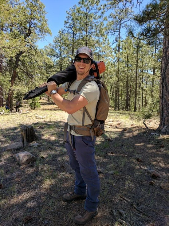 A man wearing sunglasses, a cap, and outdoor clothing, standing in a forest with tall trees, carrying a backpack and holding a camera or equipment, smiling at the camera.